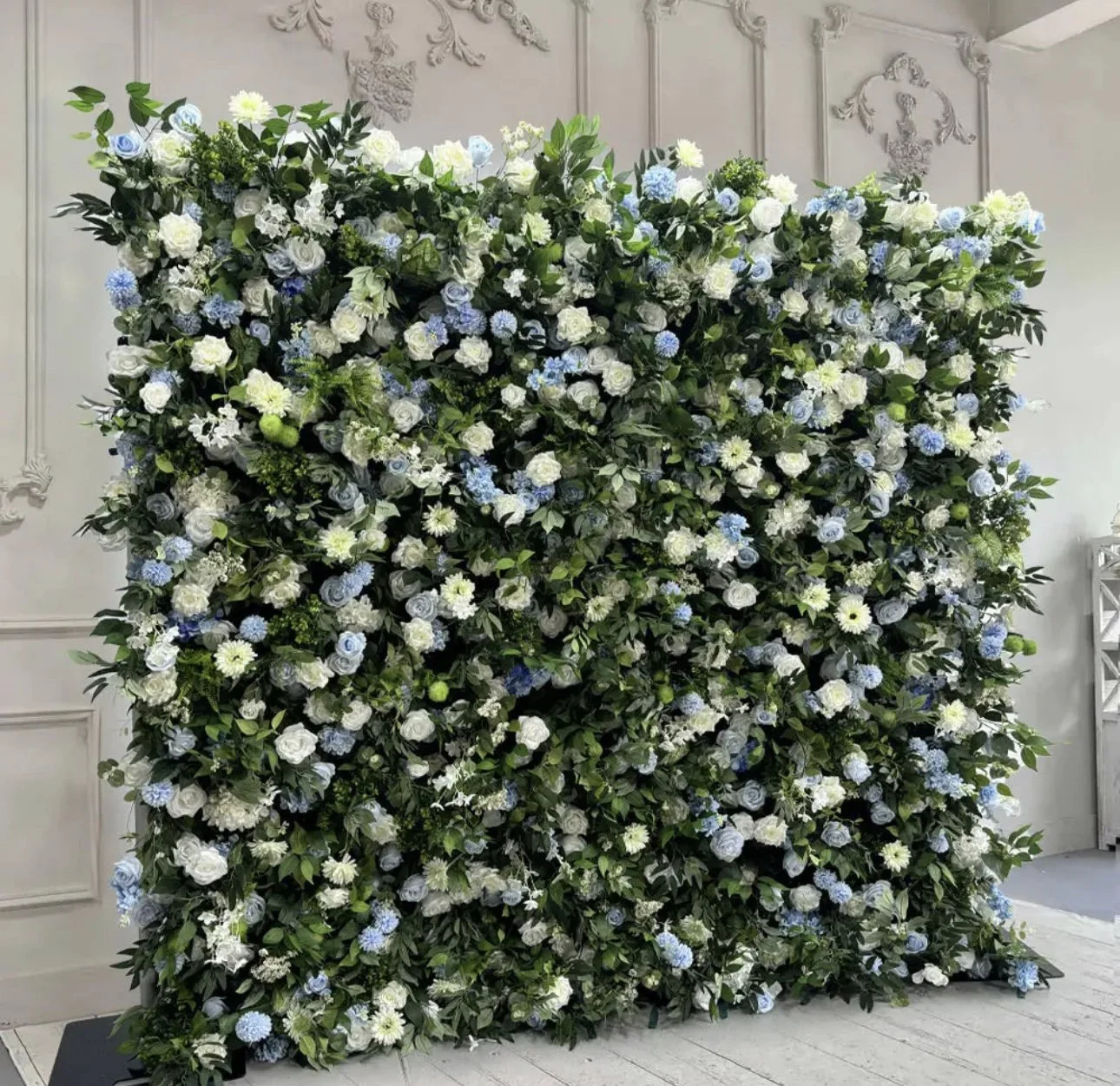 A large floral wall arrangement with white and light blue flowers and green foliage, set against a decorative, white wall with ornate molding.