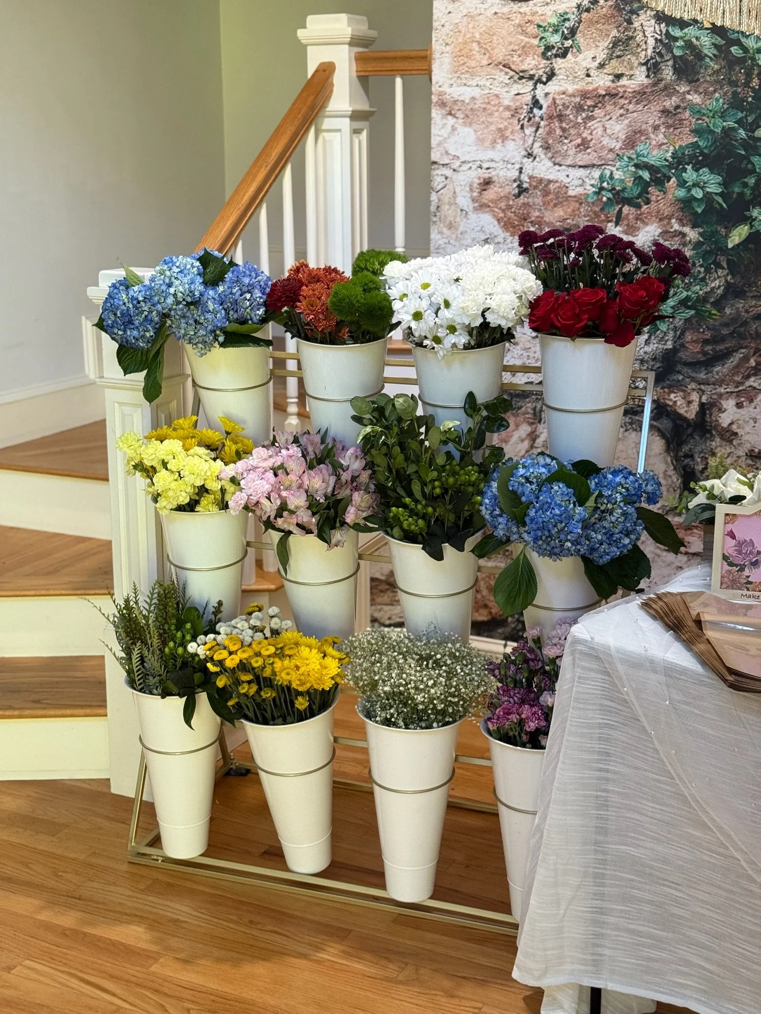 Multiple colorful flower arrangements in white vases displayed on a multi-tiered metal stand near a staircase and a stone wall background.