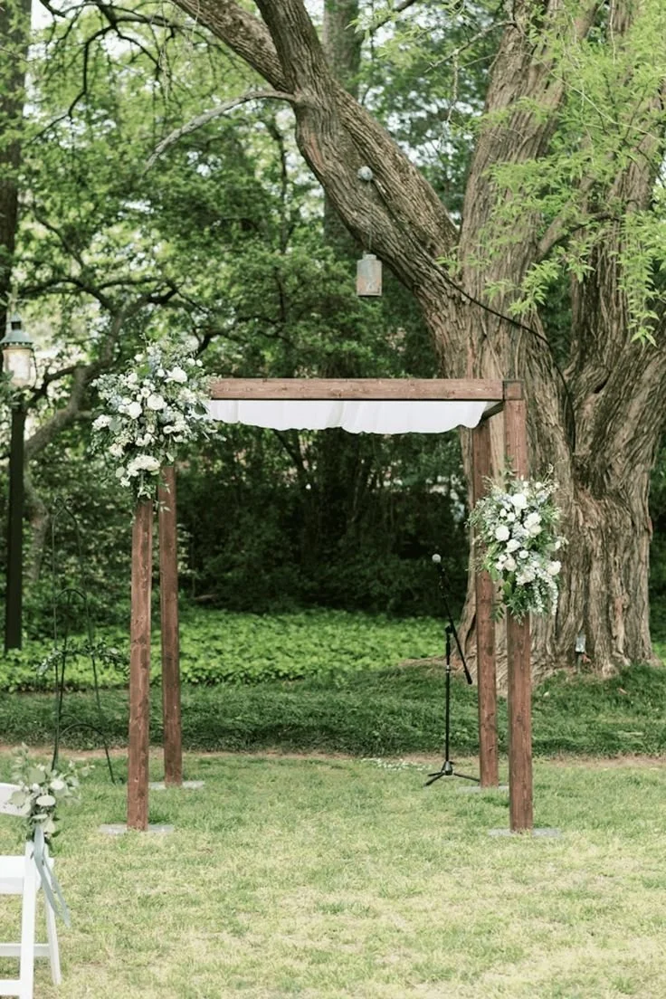 Outdoor wedding ceremony setup with a wooden arch decorated with white flowers, planted in a grassy area with trees in the background.