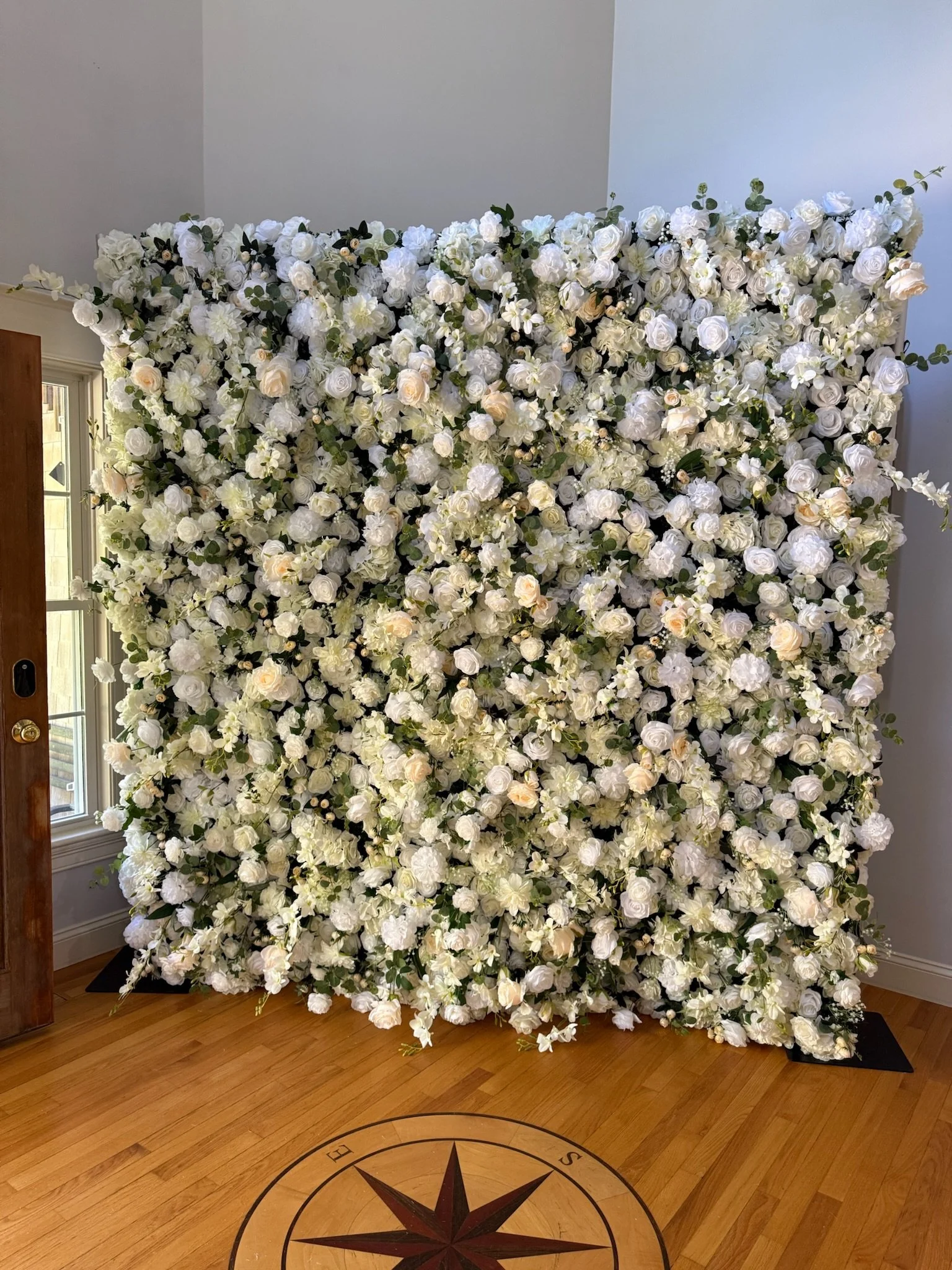 A large square floral wall adorned primarily with white roses and white flowers, with some greenery, placed indoors against a corner wall near a window and wooden floor with a compass design.