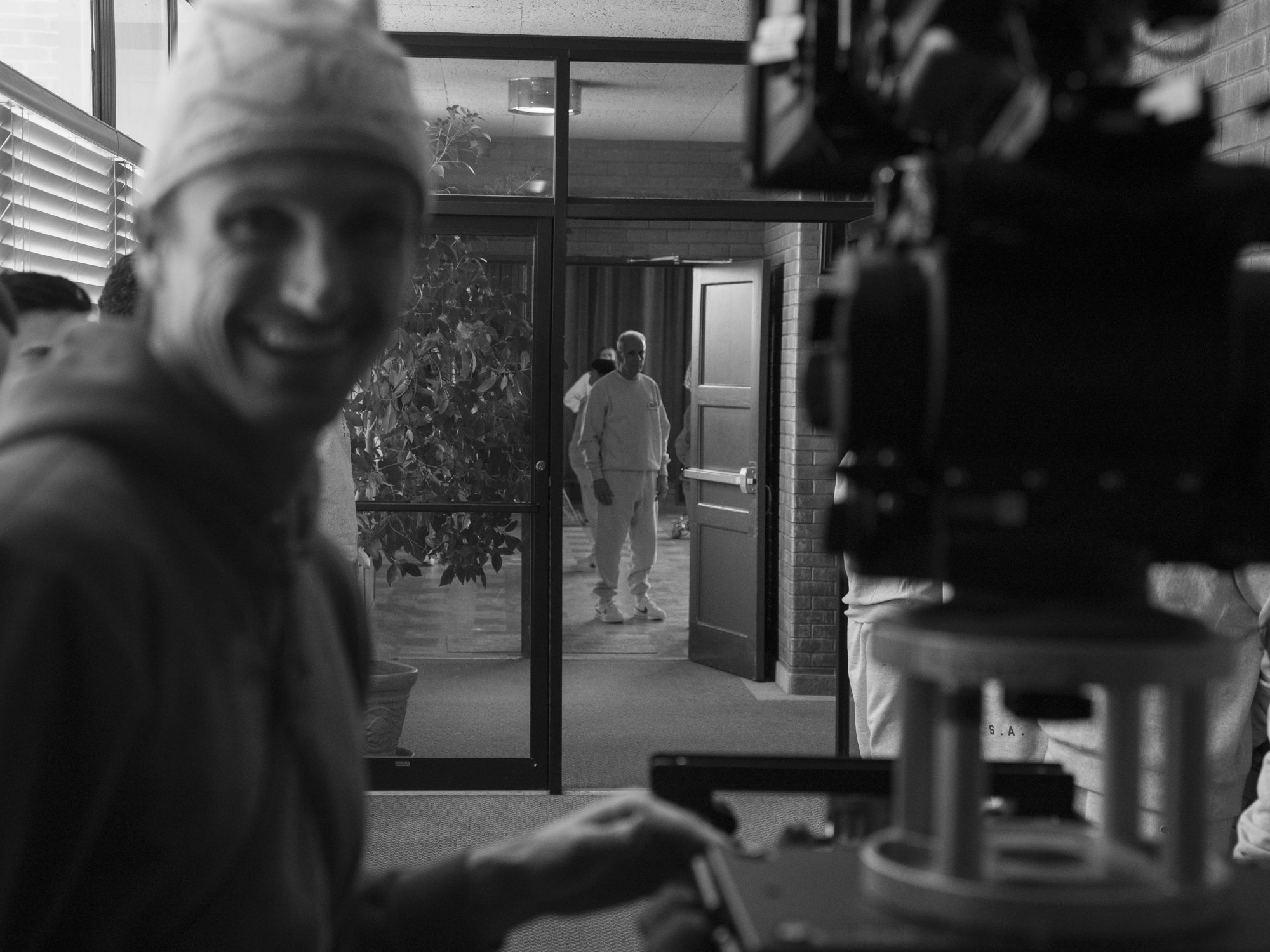 Black and white photo of a movie set featuring a close-up of a smiling person wearing a beanie, a large camera rig, and another person in the background walking through a door in a casual outfit.