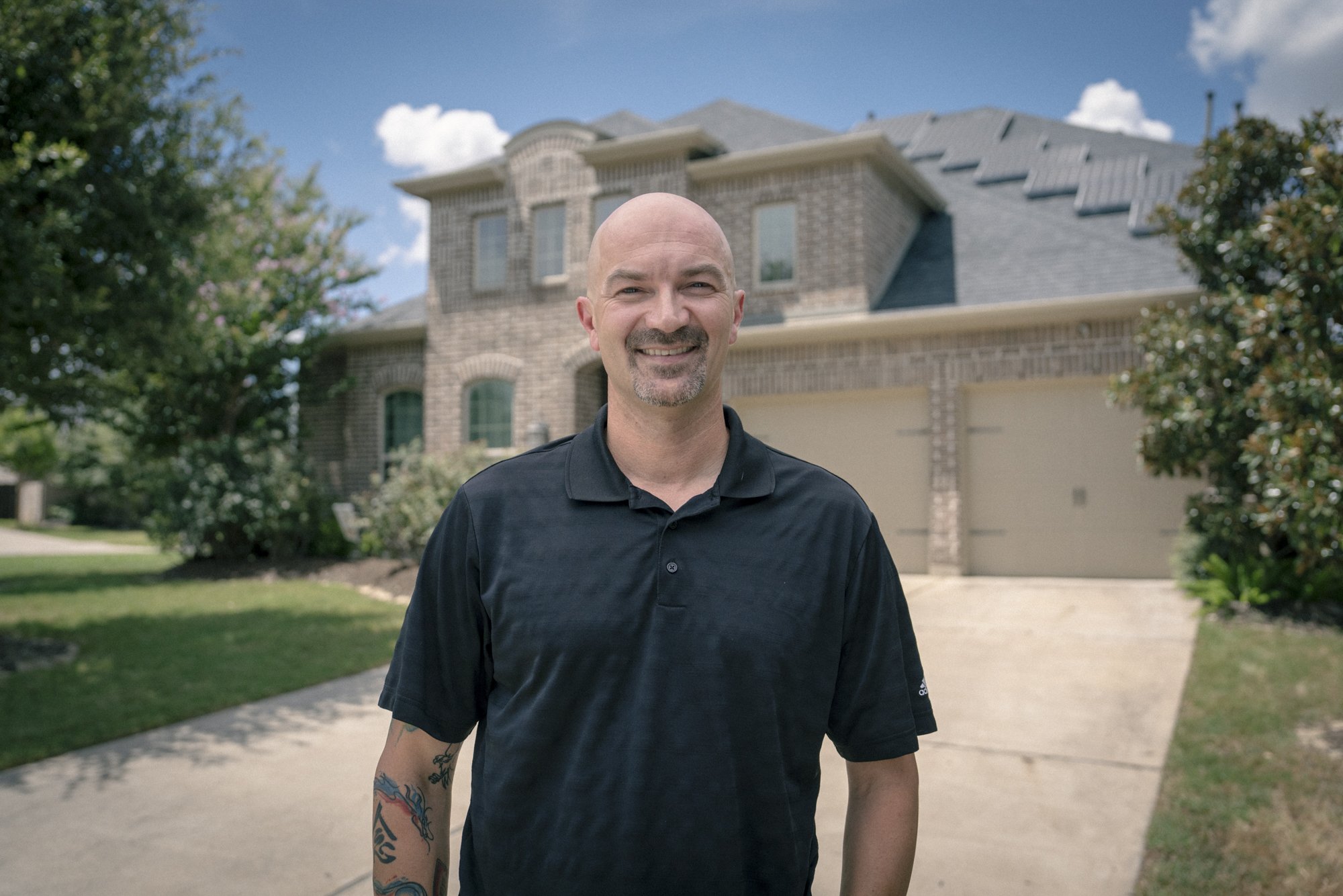 A bald man with a goatee wearing a black polo shirt stands smiling in front of a brick house with a two-car garage. There are trees and shrubs in the yard, and the sky is partly cloudy.