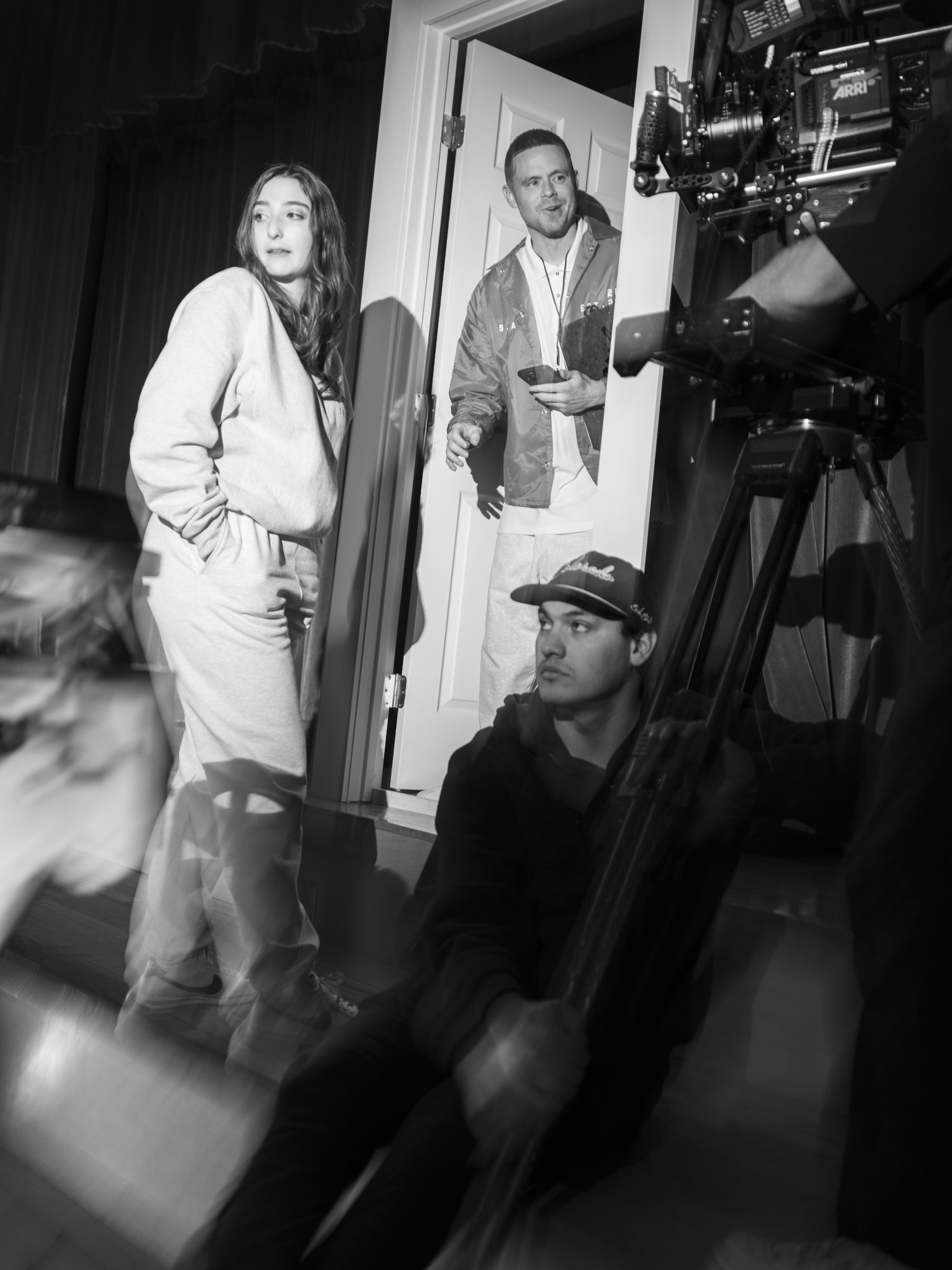 Black and white scene of a film set with a woman in casual wear and a man holding a smartphone standing near a door. A cameraman operates a large camera.