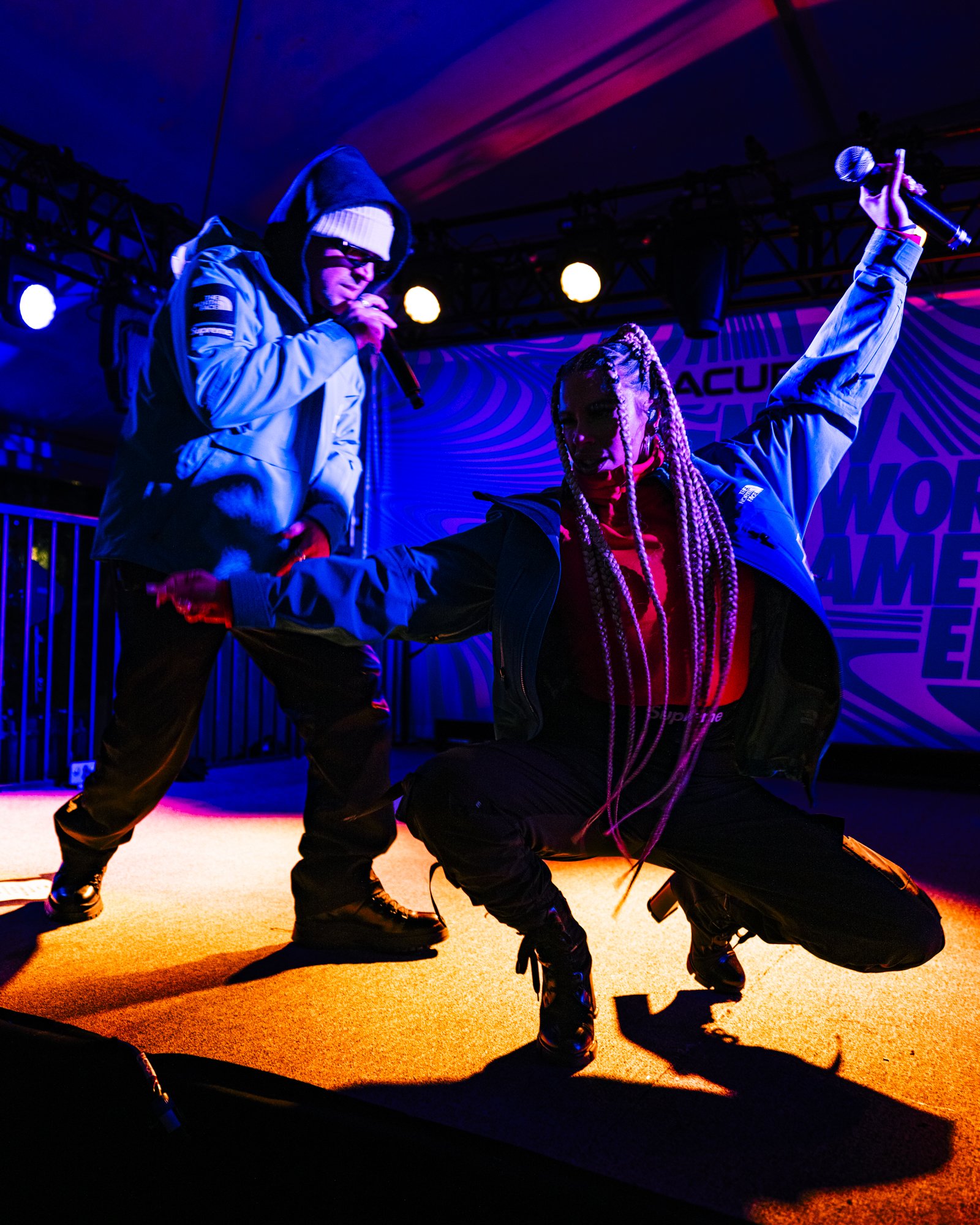 Two performers in dynamic poses on stage, one holding a microphone, both wearing stylish urban streetwear and illuminated by colorful stage lights.