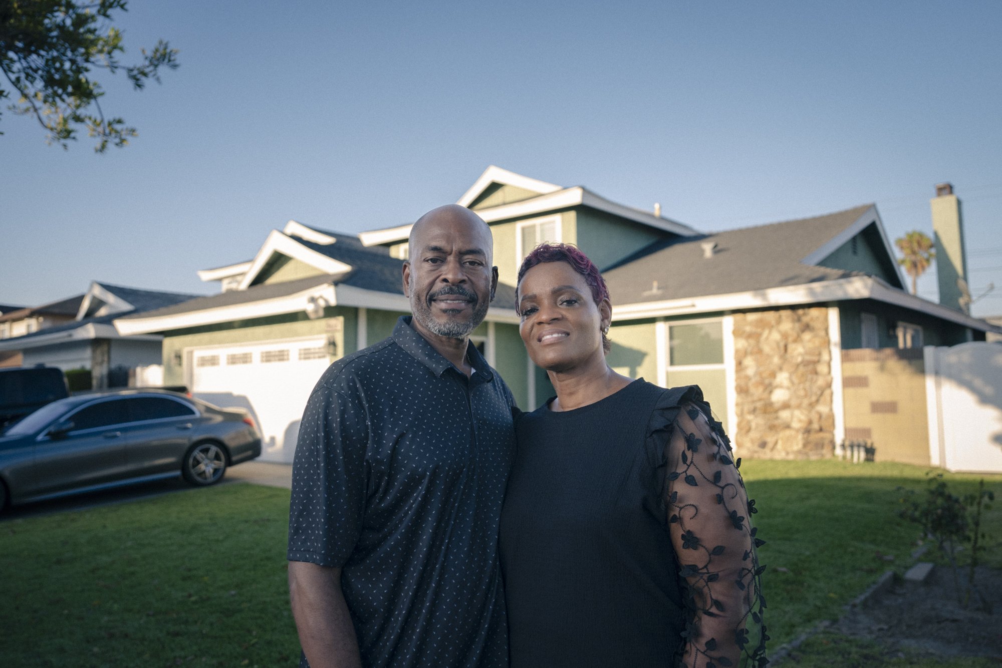 A man and woman standing in front of a house with a car parked in the driveway.