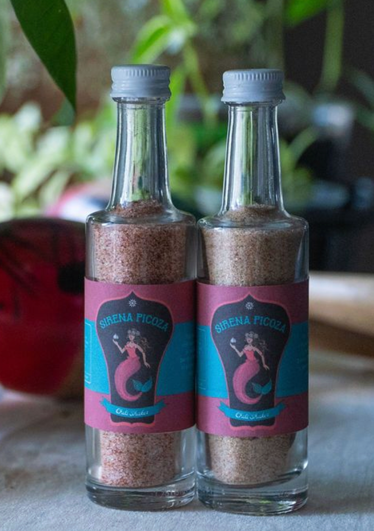 Two glass bottles filled with pink salt, each with a label featuring a mermaid and the text "Sirena Picoza" and "Old Water".