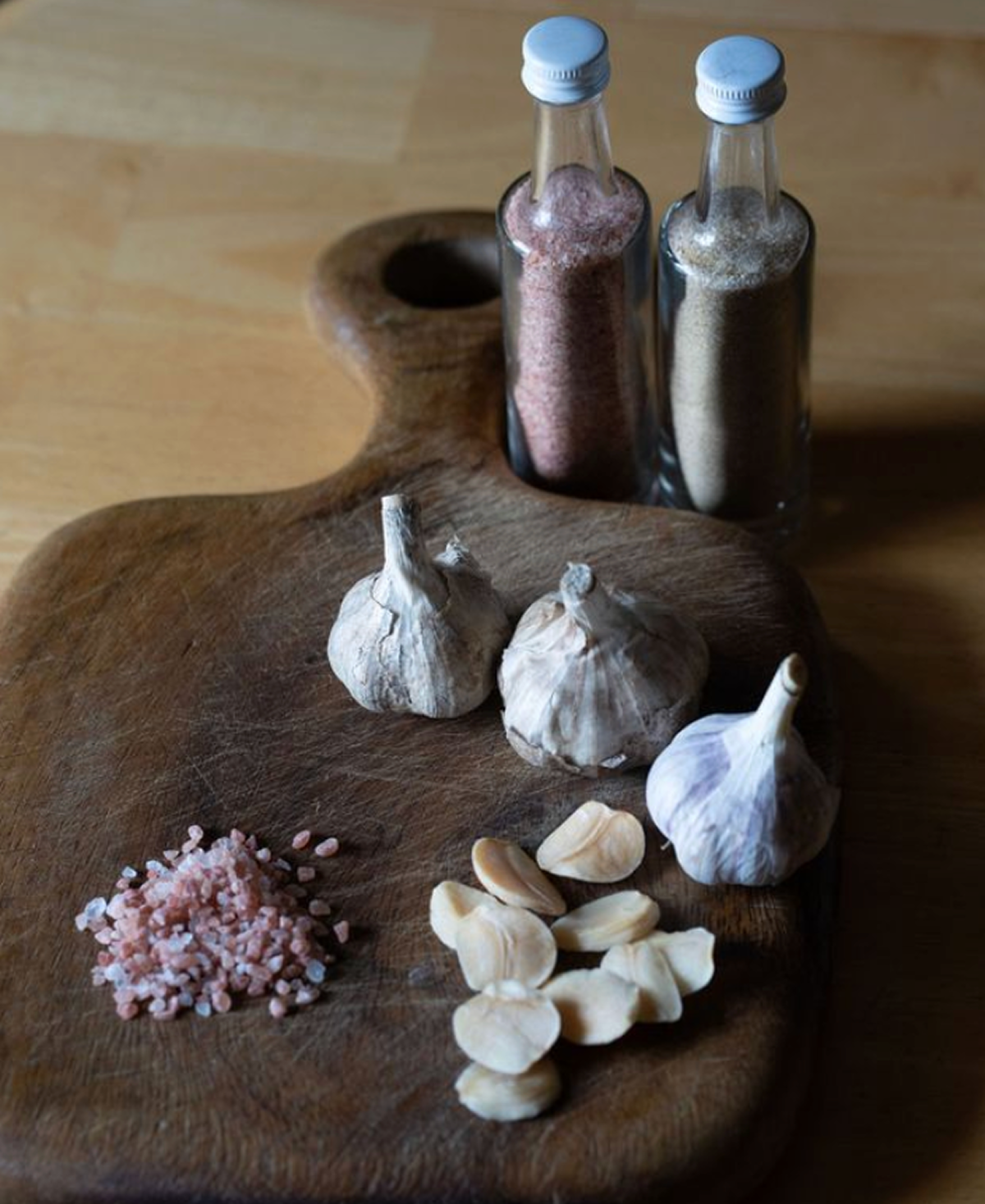 A wooden cutting board on a wooden surface with three whole garlic bulbs, garlic cloves, pink sea salt, and two glass bottles of salt and pepper.