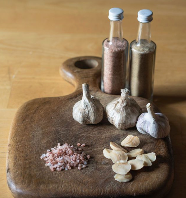 Garlic bulbs, sliced garlic cloves, pink Himalayan salt, and black pepper in grinders on a wooden cutting board.