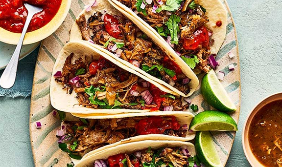 Four tacos filled with shredded meat, chopped onions, cilantro, and salsa, served on a oval plate with lime wedges and sides of salsa and sauce.