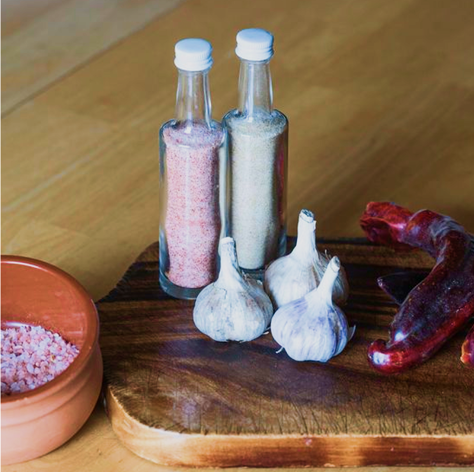 Two glass bottles filled with pink and white granular substances are on a wooden surface, with garlic bulbs in front and a reddish clay chili pepper to the side.
