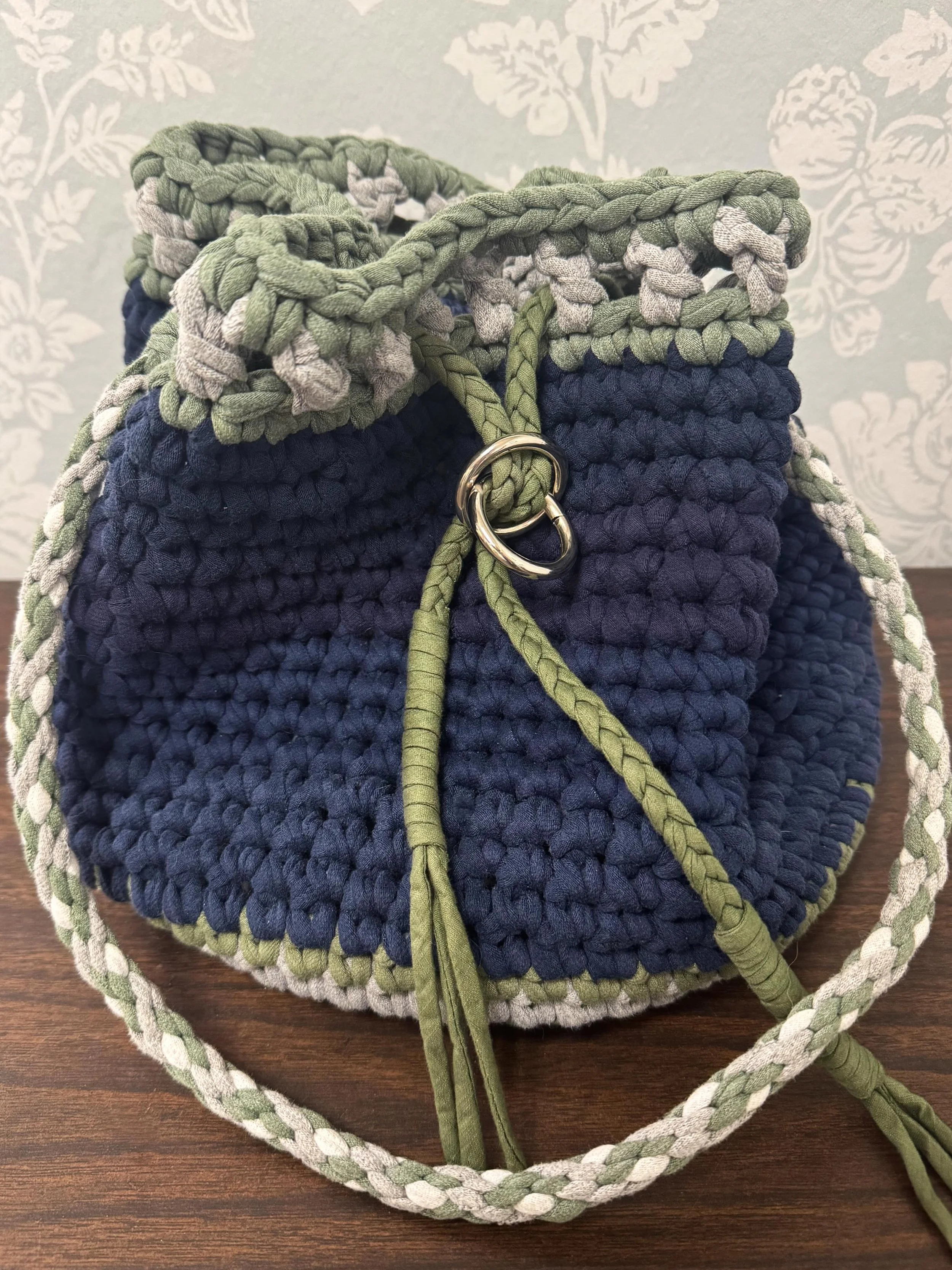 This handcrafted bag was crocheted by one of our volunteers using ten repurposed t-shirts. The metal hardware was salvaged from a damaged handbag, the lining was created from dress shirts, and the cord was originally a decorative dress belt. Every ma