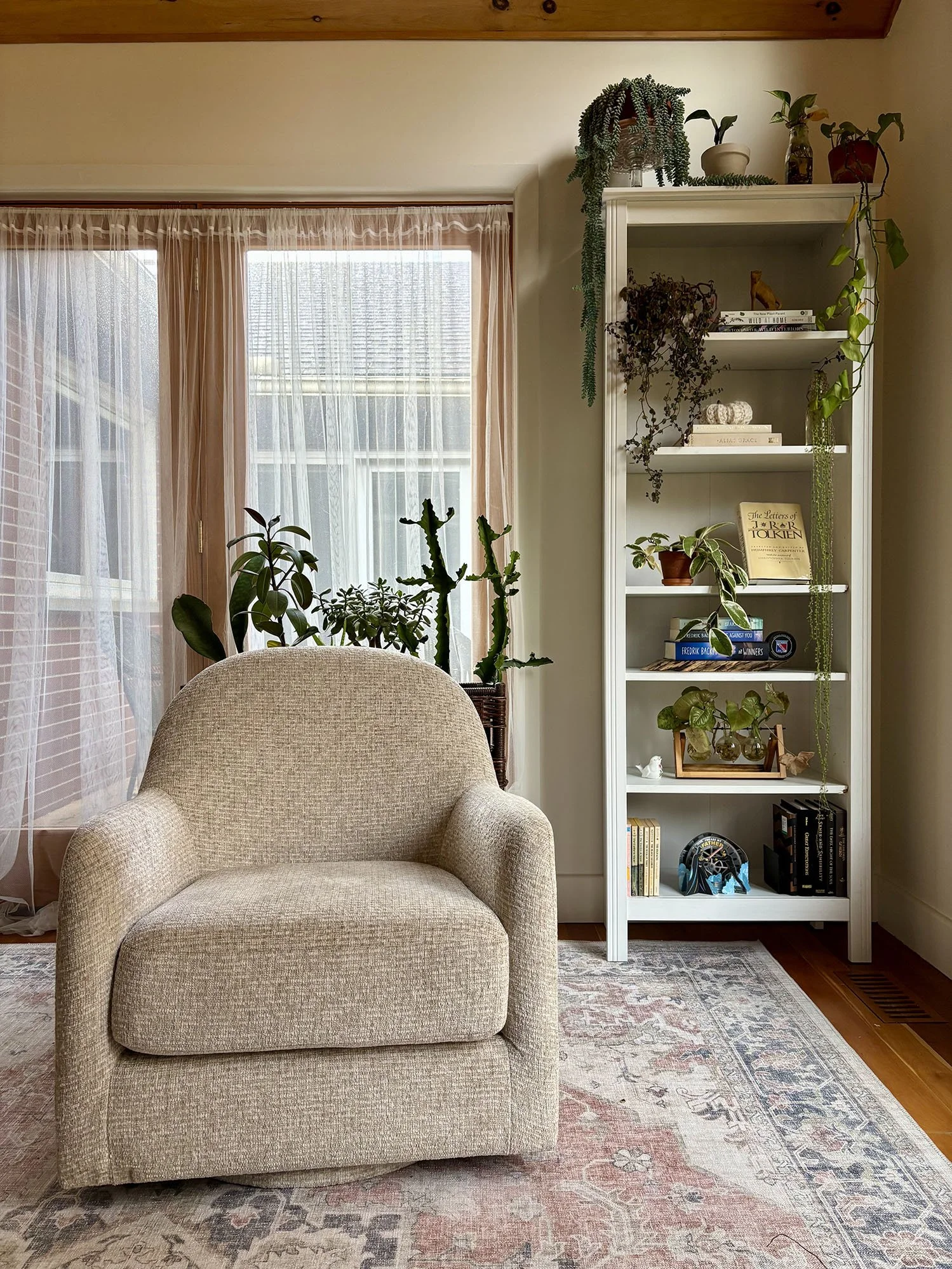 A cozy psychotherapist’s office with a soft beige armchair centred on a patterned rug, surrounded by houseplants near sheer-curtained windows and a tall white bookshelf filled with books and greenery.
