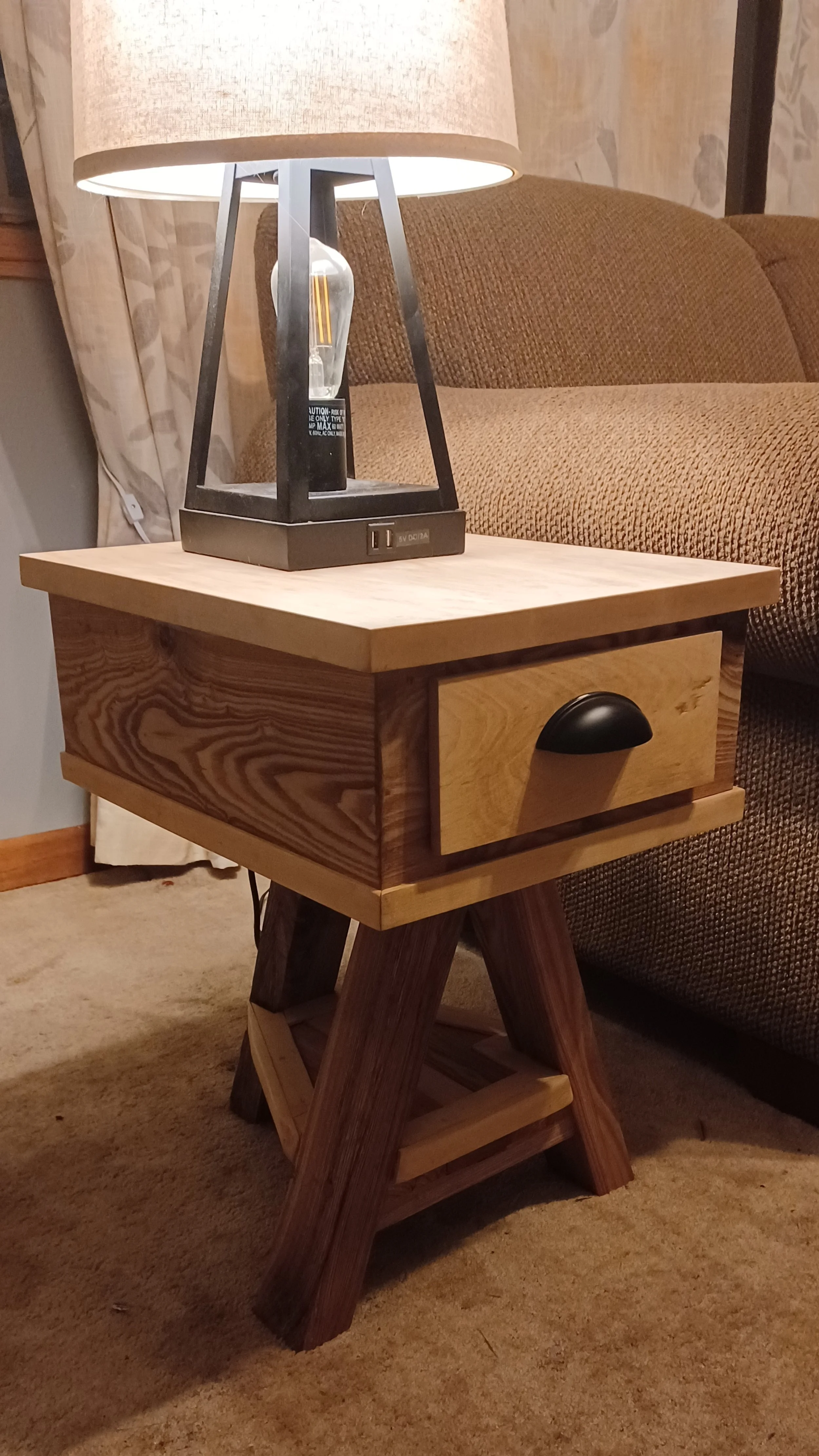 A wooden side table with a drawer and a lower shelf next to a brown sofa, with a lamp on top of the table. The lamp combines a beige lampshade with a black metal frame and an Edison-style bulb. The background has a curtain and a wall with a stone pat