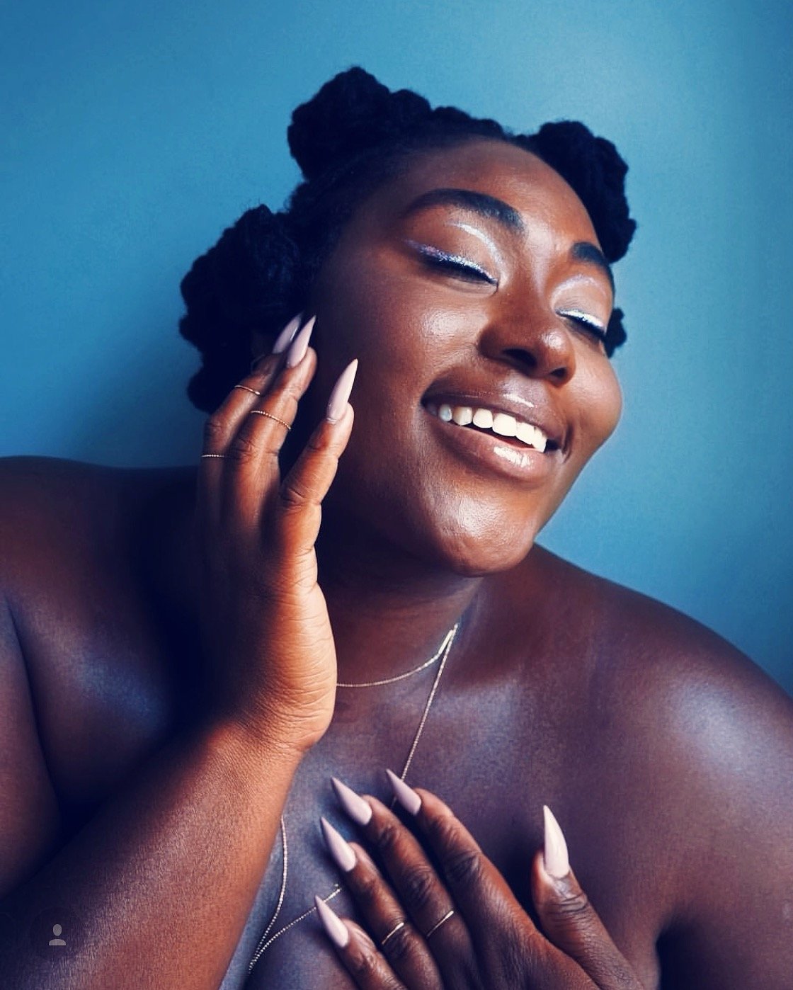 A smiling woman with dark skin, styled hair in twists, wearing makeup, jewelry, and long nails, posing against a blue background.
