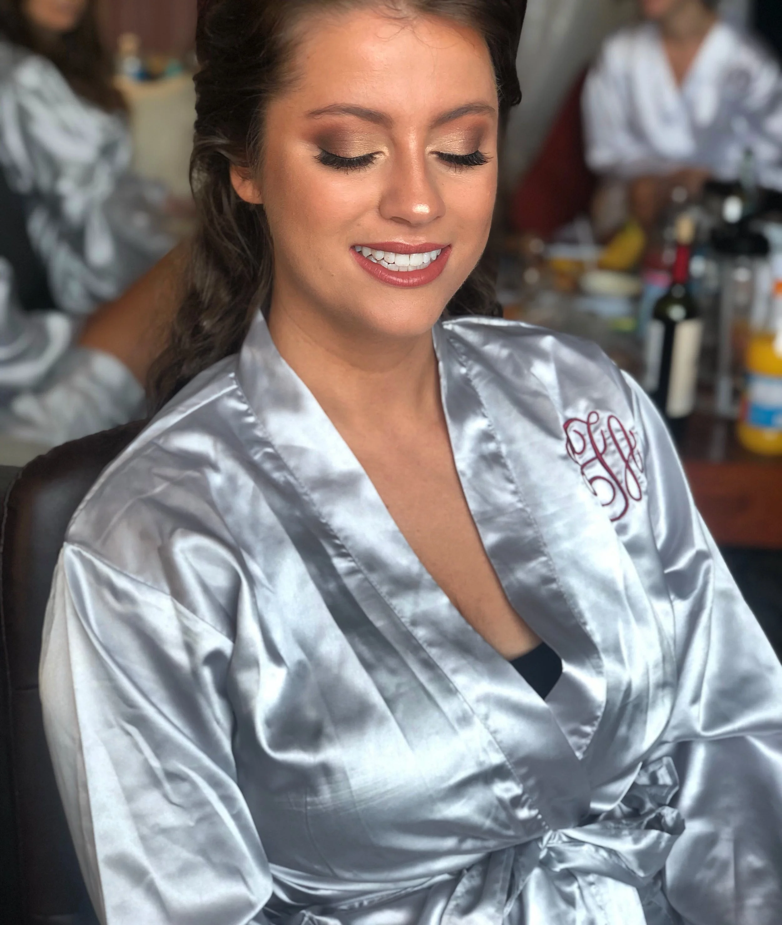A woman in a silver satin robe with a monogrammed embroidery, smiling with her eyes closed, in a dressing room with makeup and hair supplies in the background.