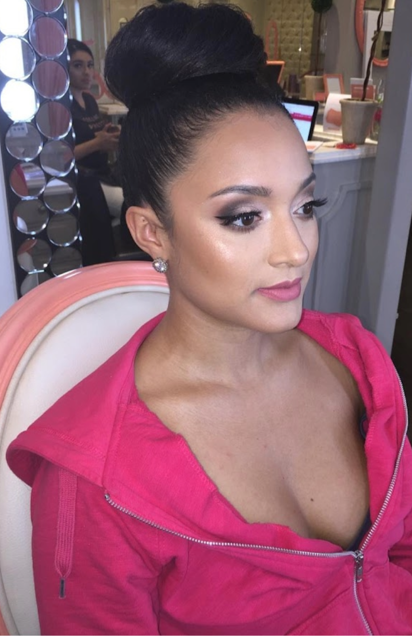 A woman with dark hair styled in an elegant updo, wearing makeup including eyeliner and lipstick, dressed in a pink zip-up hoodie, sitting on a white chair in a salon or studio setting.