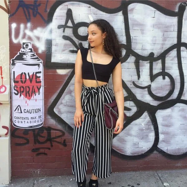 A young woman with curly hair, wearing a black crop top and high-waisted black and white striped pants, stands in front of graffiti-covered brick wall holding a maroon handbag.