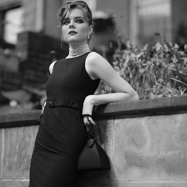 A woman in a sleeveless black dress and lace choker stands outdoors, leaning on a concrete wall, holding a black purse, with a blurred urban background.