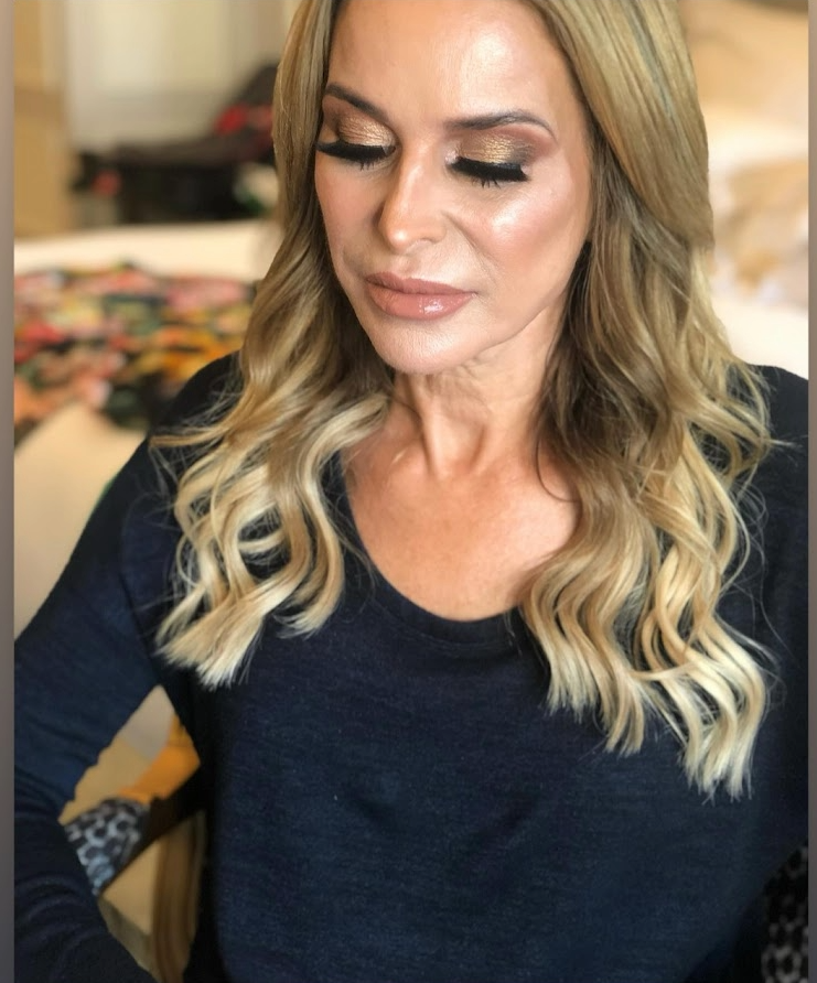 A woman with long blonde wavy hair, wearing makeup with glittery eyeshadow and glossy lips, sitting indoors with her eyes closed.