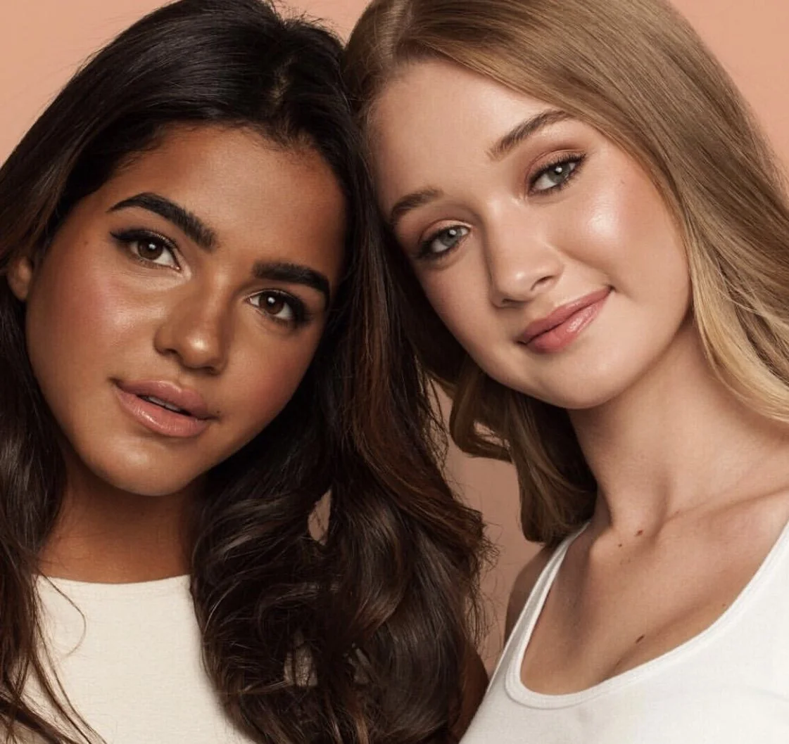 Close-up of two young women smiling with their heads close together, one with dark hair and the other with light hair, against a plain background.