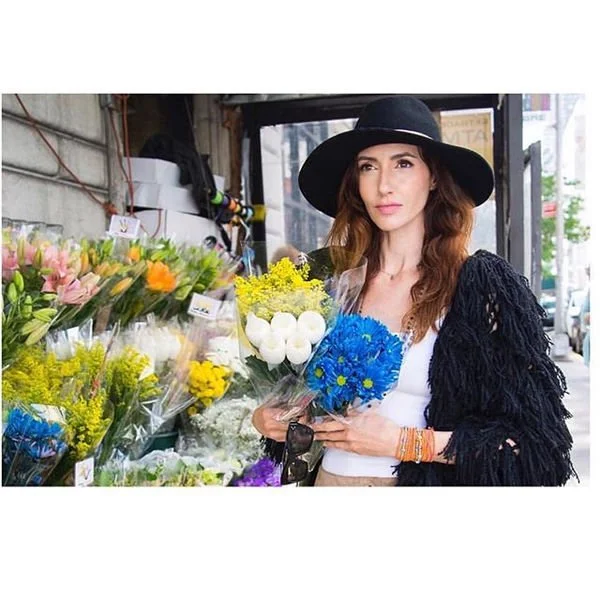 Woman shopping for flowers at an outdoor market, holding a bouquet of blue and white flowers, wearing a wide-brimmed black hat and a black fringed jacket.