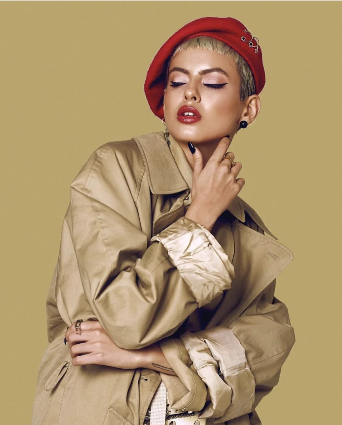 A woman with short blonde hair wearing a red beret, tan trench coat, and bold makeup, posing with her eyes closed against a beige background.