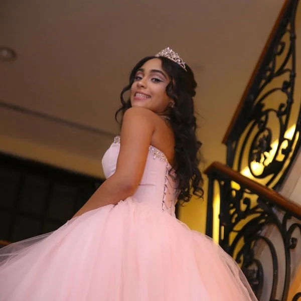 Young woman in a pink ball gown with a tiara, smiling and looking over her shoulder indoors near a staircase with ornate black banister.