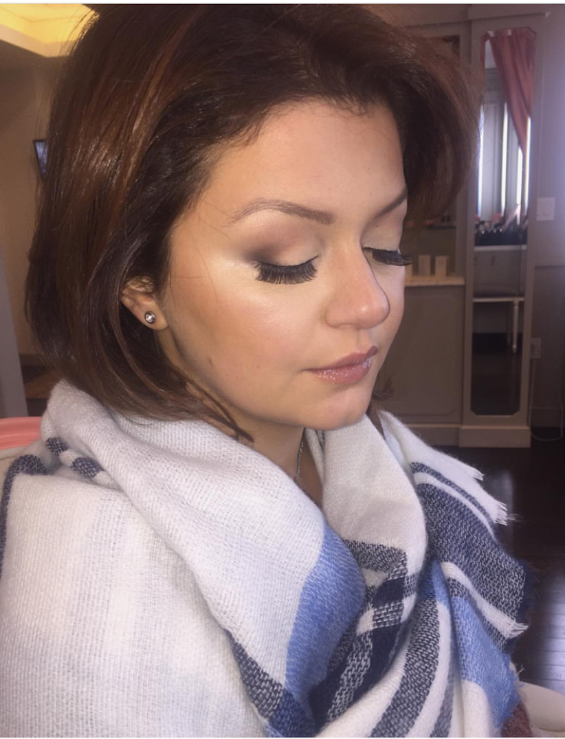 A woman with short brown hair and makeup, wearing earrings and a white shawl with blue and black plaid pattern, sitting with eyes closed in a room with pink curtains and a desk in the background.