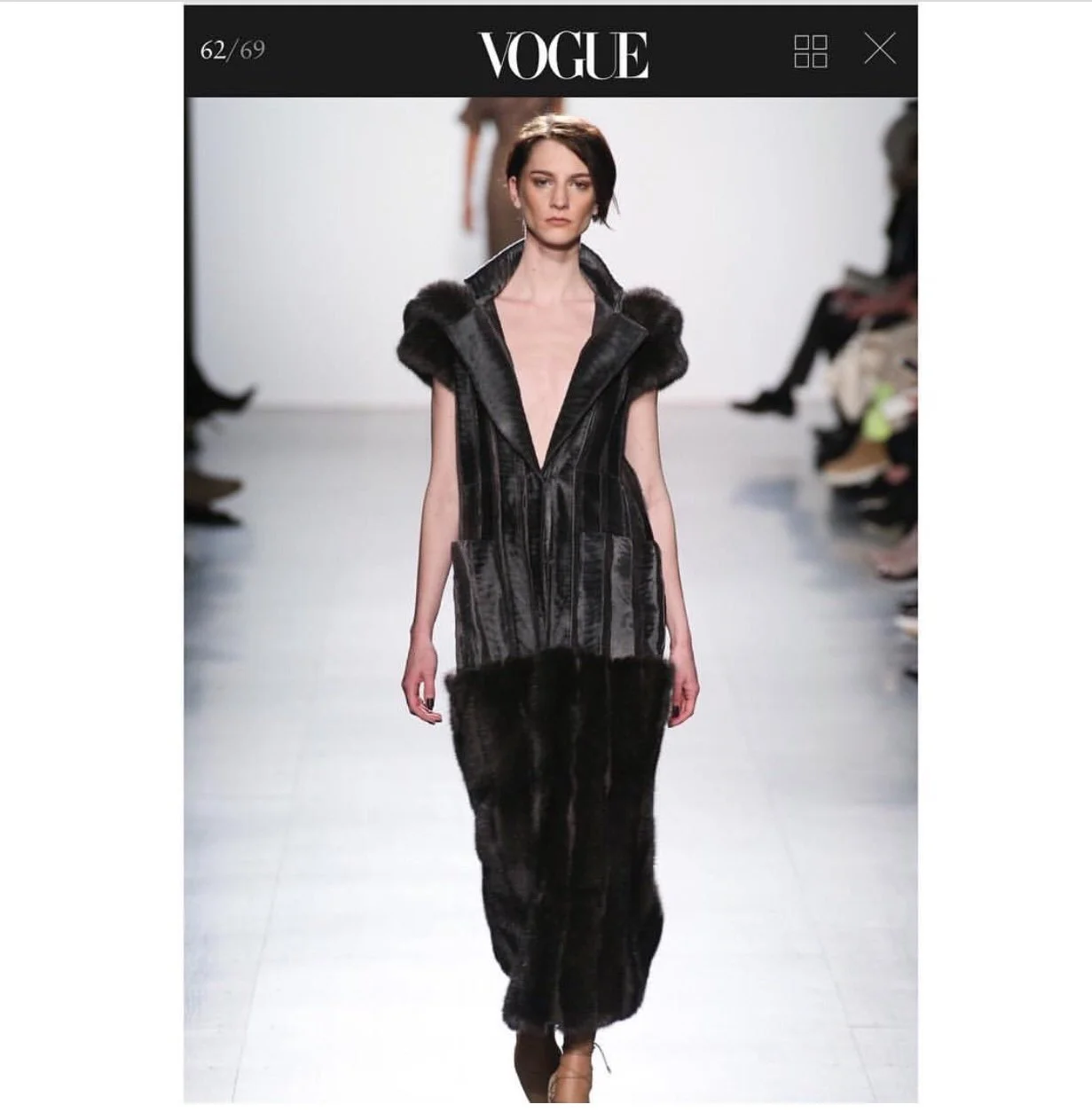 Model walking on runway during a fashion show, wearing a black dress with fur accents at the shoulders and hem, under bright lights with audience seated on either side.