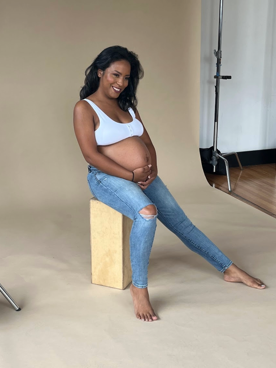 A pregnant woman with curly black hair sitting on a wooden block in a photography studio, smiling and looking down, barefoot, wearing a white tank top and ripped blue jeans.