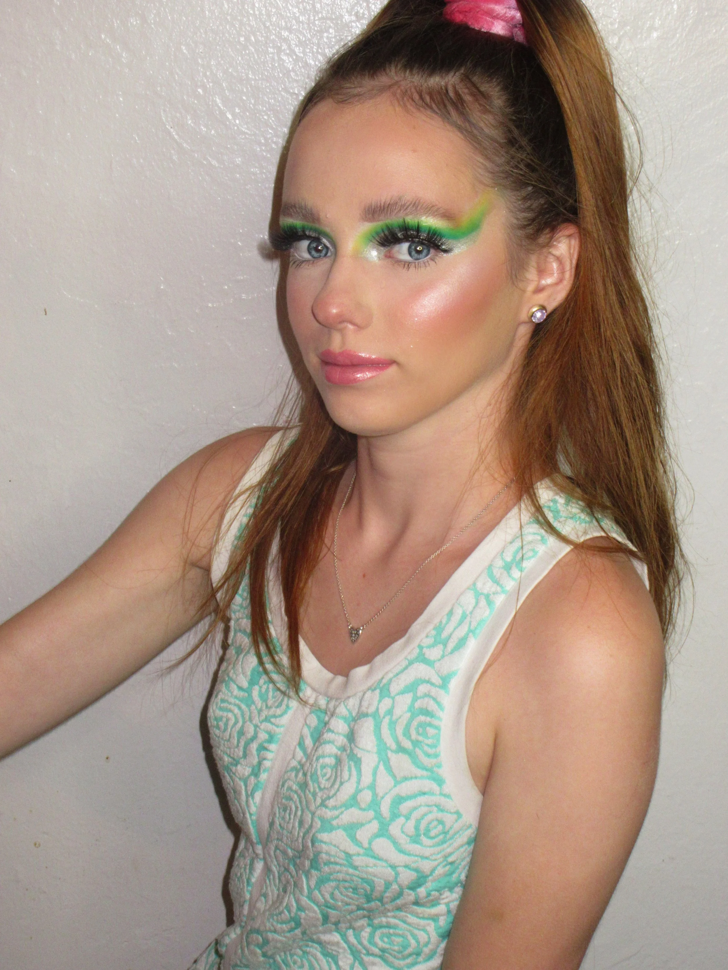 A young woman with vibrant green and yellow eyeshadow, long lashes, and shiny pink lips. She has straight, reddish-brown hair tied up in a high ponytail with a pink scrunchie, wearing a sleeveless white top with turquoise swirl patterns and a silver 