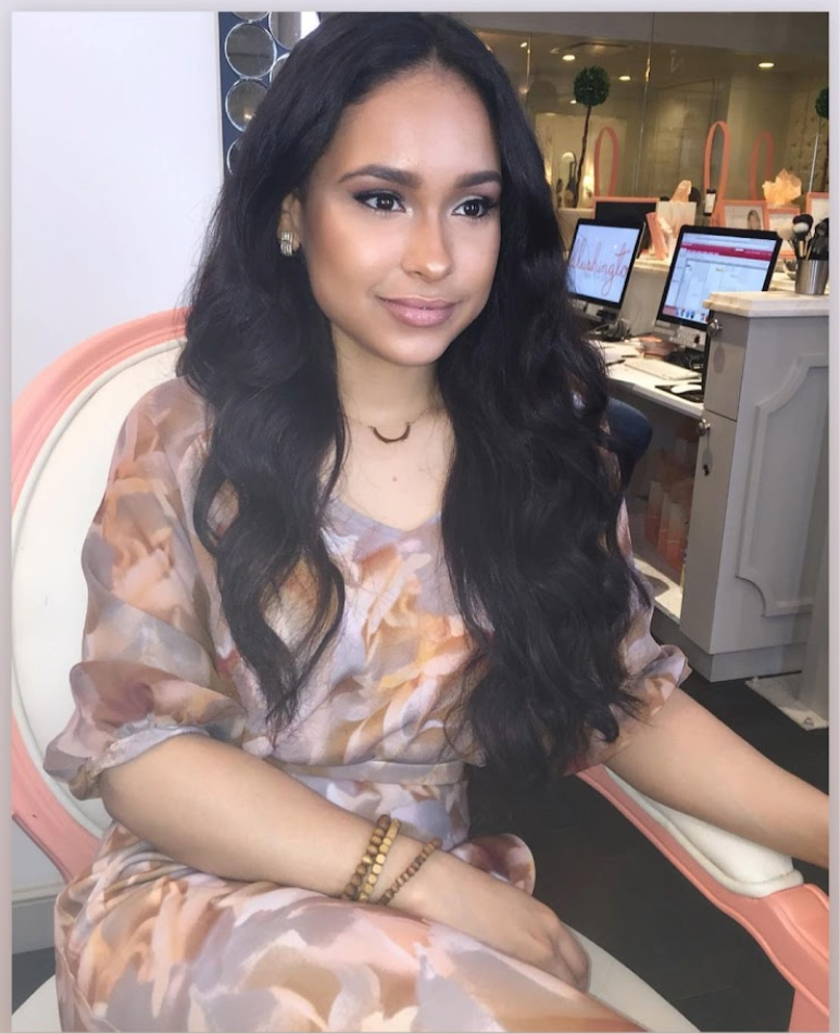 A woman with long, wavy black hair and makeup sitting on a pink and white chair. She wears a floral dress, jewelry, and is in an indoor setting with computer monitors and makeup items in the background.
