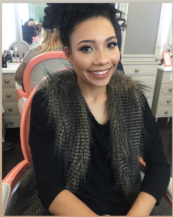 Smiling woman with curly hair sitting in a salon chair with a cape that has faux fur trim, in a hair salon with mirrors and salon supplies in the background.