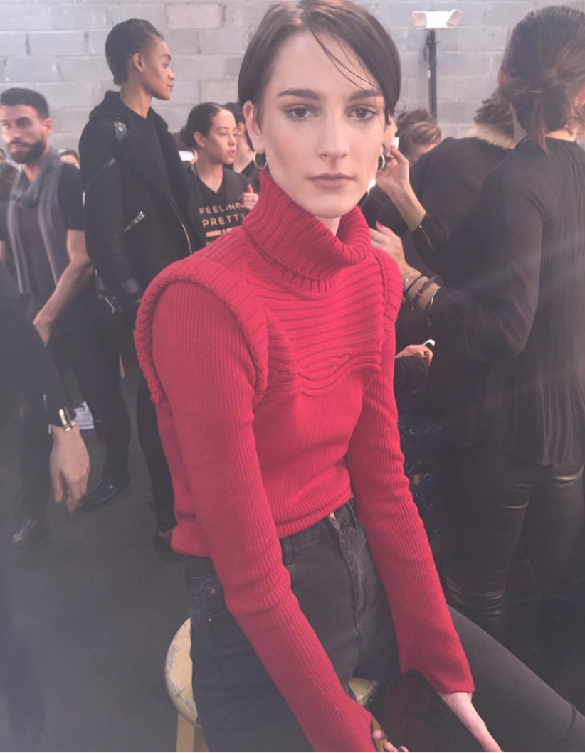A young woman with short dark hair and hoop earrings, wearing a red turtleneck sweater, sitting on a stool in a busy backstage area with a gray brick wall, surrounded by people preparing for an event.