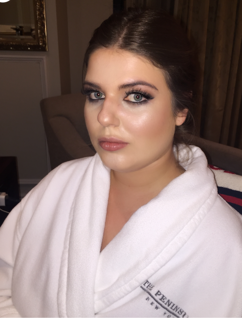 A woman with makeup, blue eyes, and dark hair in a white bathrobe sitting in a room with brown curtains and a beige chair.