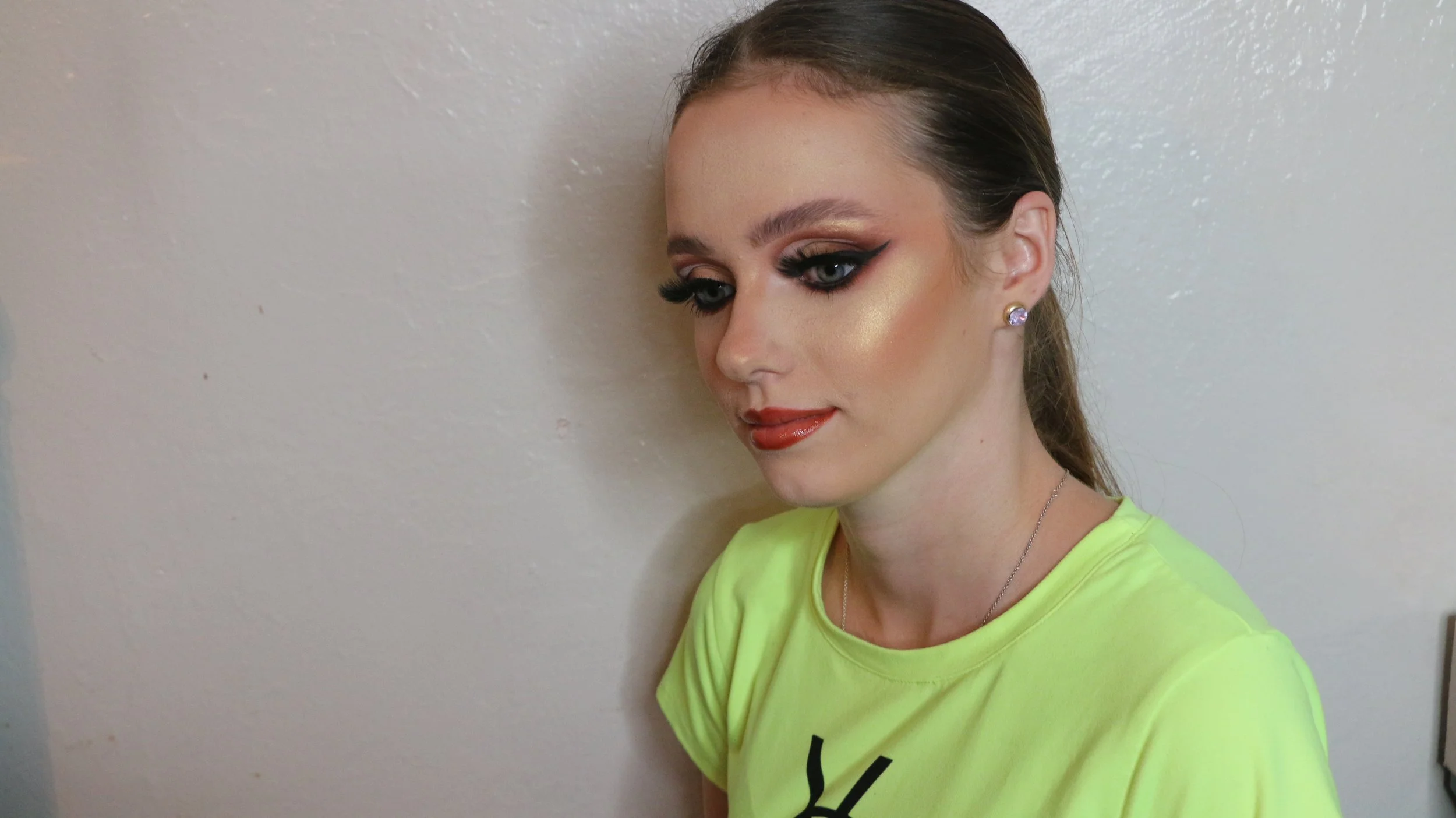 A woman with dramatic eye makeup, including winged eyeliner and false eyelashes, has highlighted cheekbones and glossy lips. She is wearing a yellow T-shirt and a silver necklace, positioned against a plain white wall.