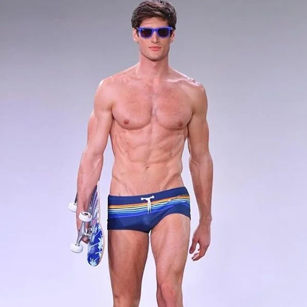 Male model wearing blue swim trunks with yellow and white stripes, blue sunglasses, holding skateboard, walking on runway with gray background.