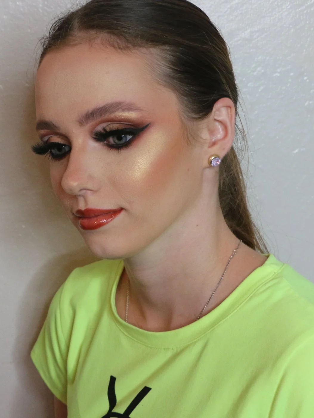 A woman with styled hair, dramatic makeup including winged eyeliner and gold eyeshadow, wearing earrings and a neon yellow shirt.