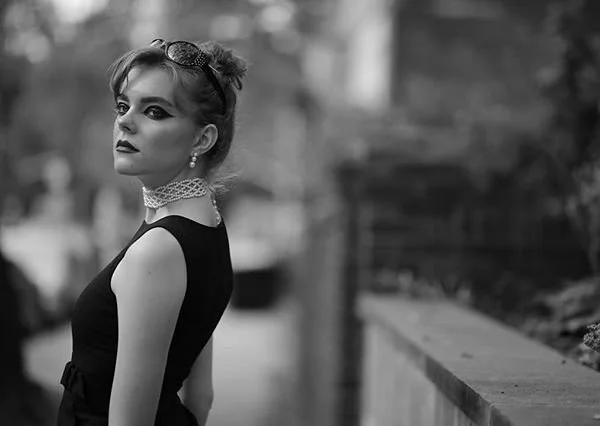 A woman with styled hair and makeup, wearing sunglasses on her head, earrings, a lace choker, and a sleeveless black top, standing outdoors near a railing in black and white.