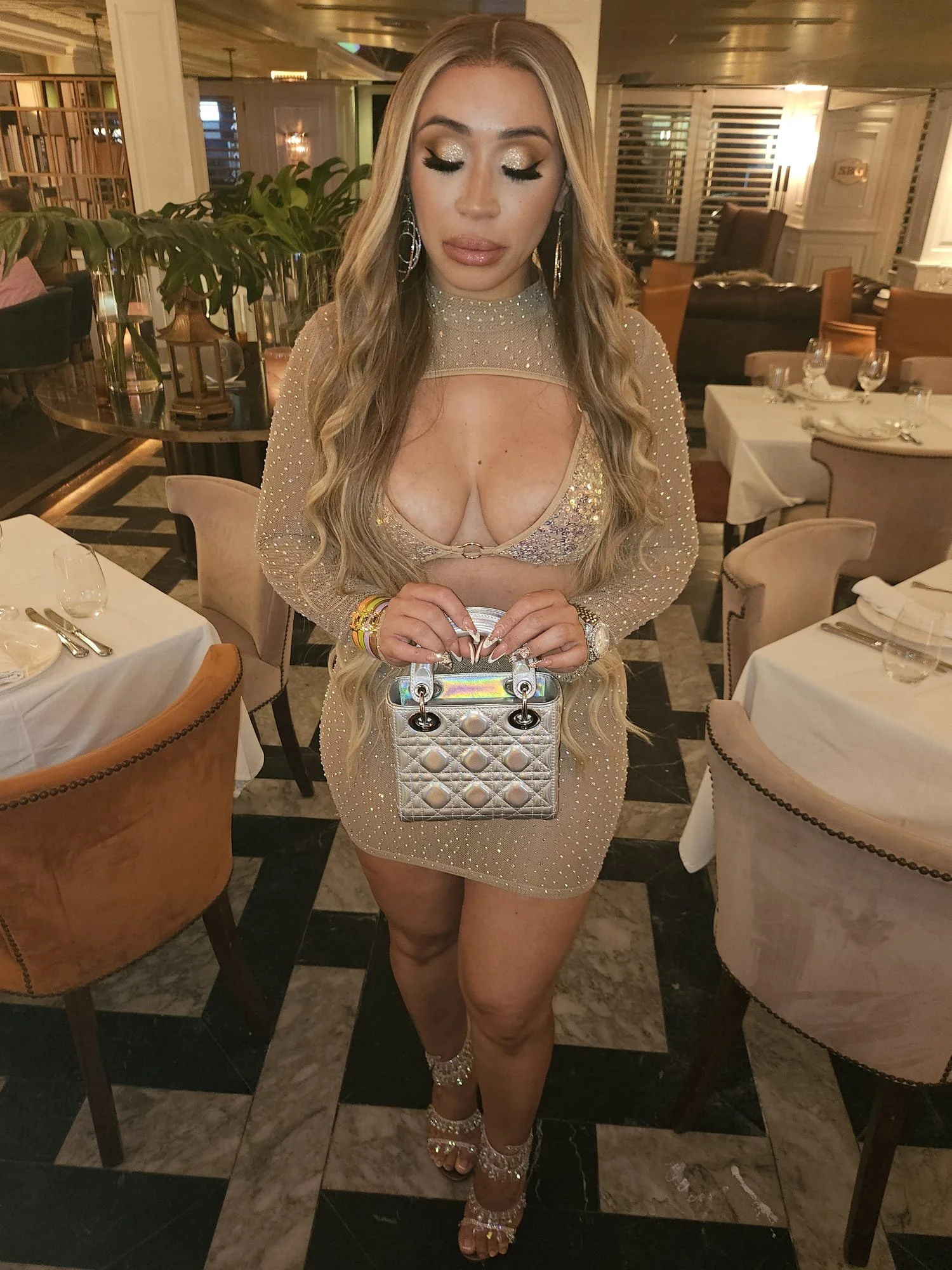 A woman with long, wavy blonde hair, applying makeup, wearing a beige, sparkling dress with a deep neckline, holding a silver handbag, in a fancy restaurant setting with white tablecloths and elegant decor.