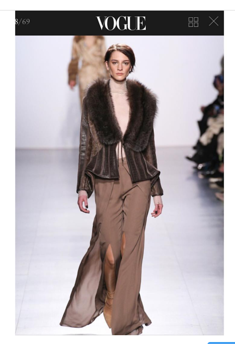 Model walking on runway wearing brown wide-leg pants with front slit, patterned top, and brown fur-collared jacket during Vogue fashion show.
