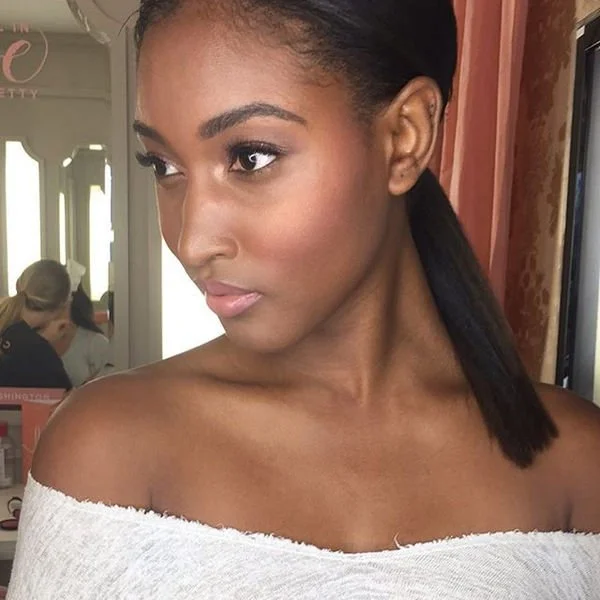 A woman with a sleek ponytail hairstyle, wearing a white off-shoulder top, with makeup and a neutral expression, in a beauty salon or similar setting.