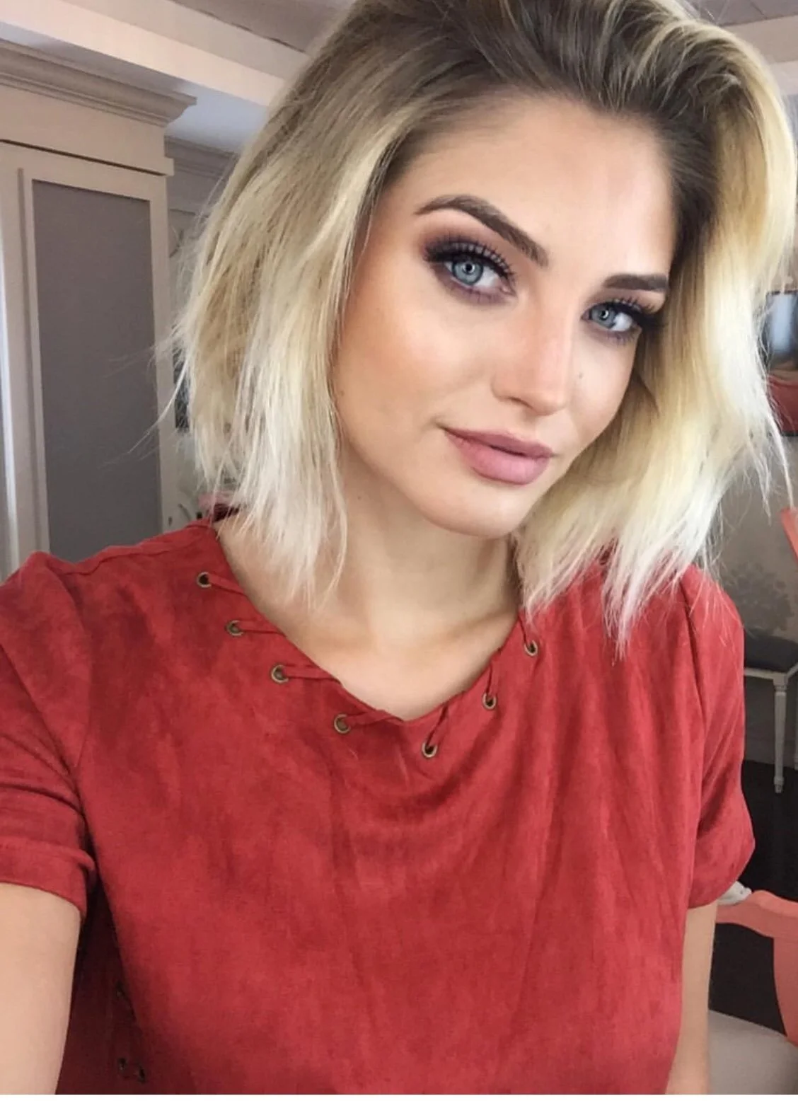 A young woman with blonde hair, blue eyes, and makeup, wearing a red shirt with eyelet details, smiling slightly, in an indoor setting.