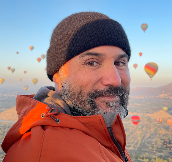 Man in orange jacket and black beanie with hot air balloons in the sky