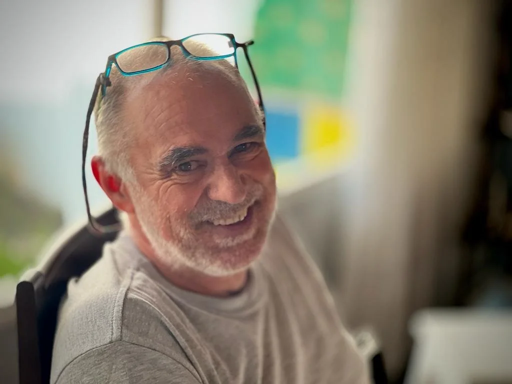 Smiling older man with glasses on his head, wearing a gray shirt.
