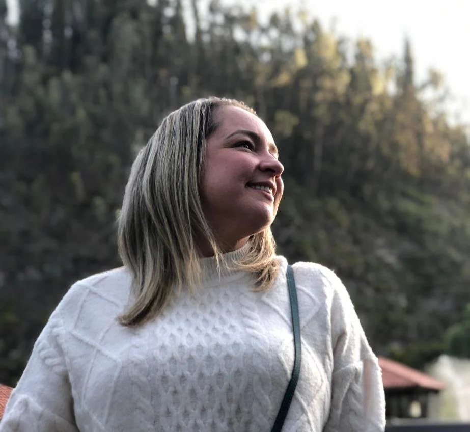 Woman in white sweater smiling outdoors with blurred forest background.