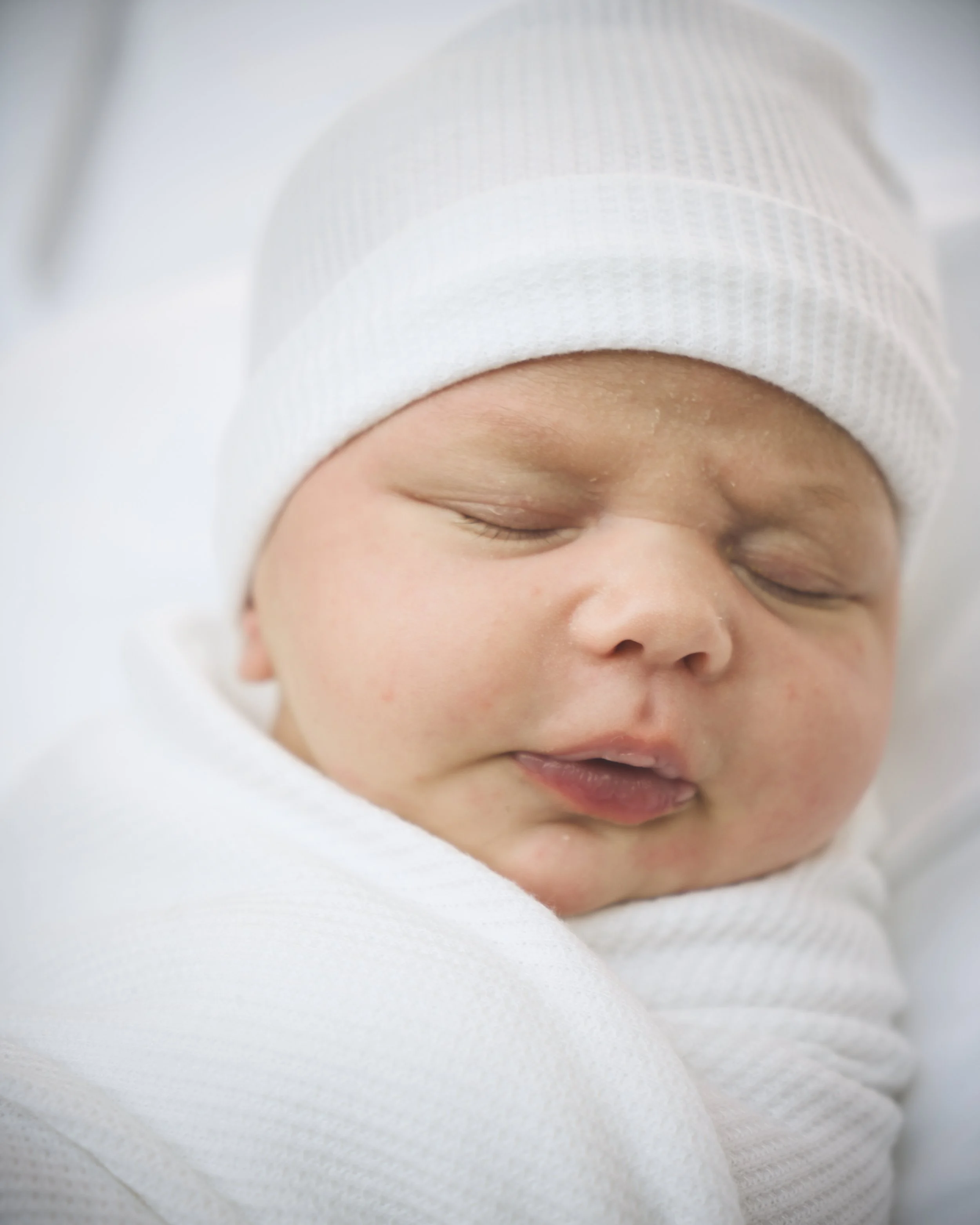 Close-up of a sleeping baby wearing a white cap and white clothing.