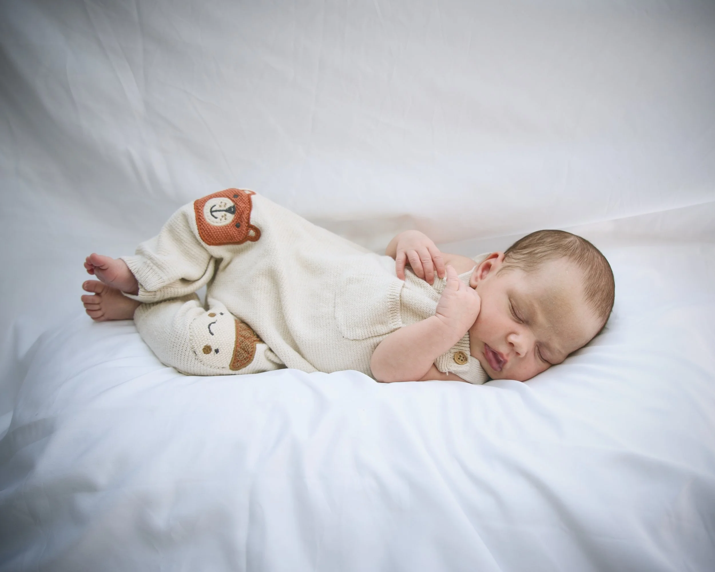 A baby lying on a white bed, sleeping peacefully, wearing a cream-colored onesie with bear patches.