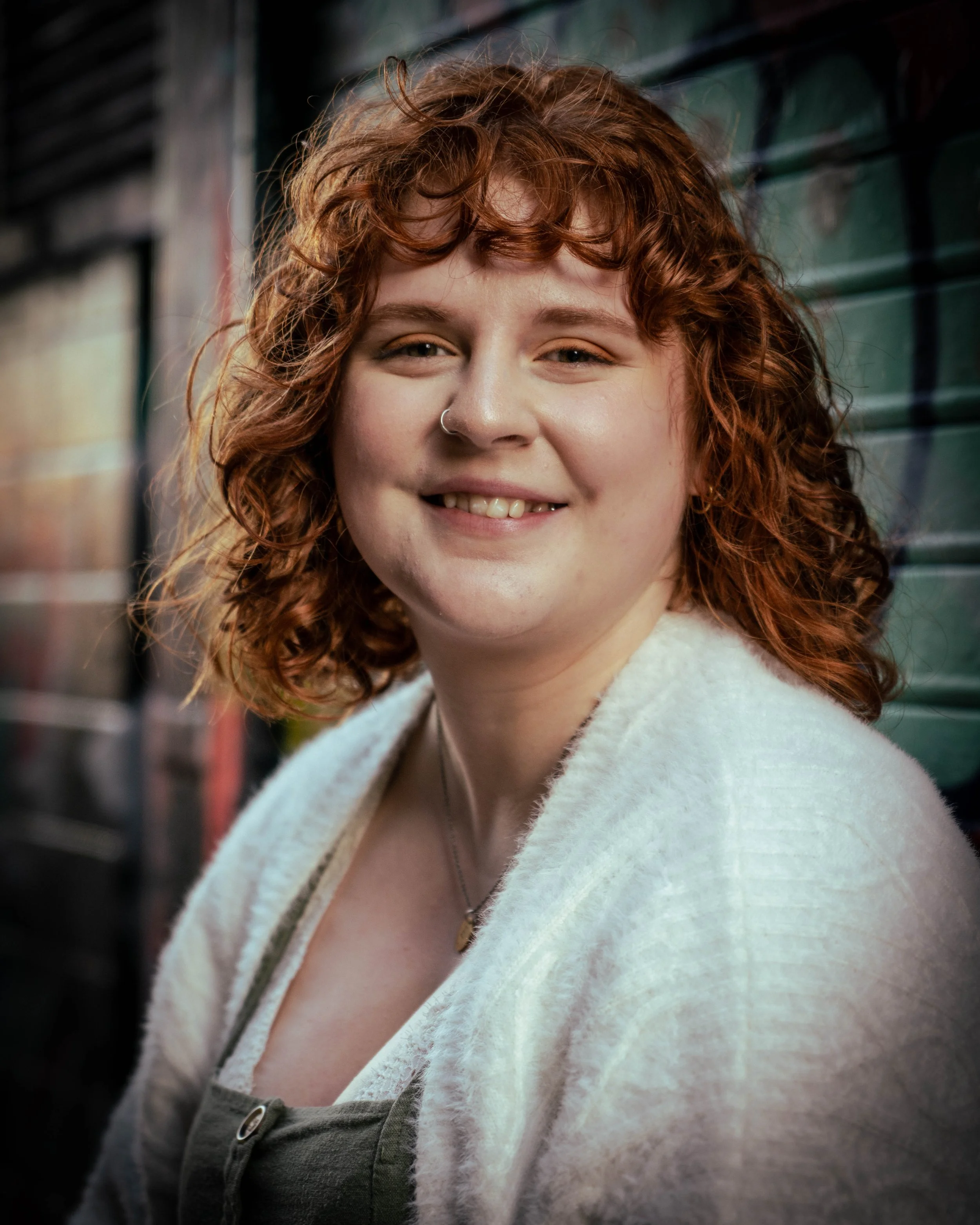A smiling young woman with curly red hair and a nose ring, standing outdoors against a weathered green wall.