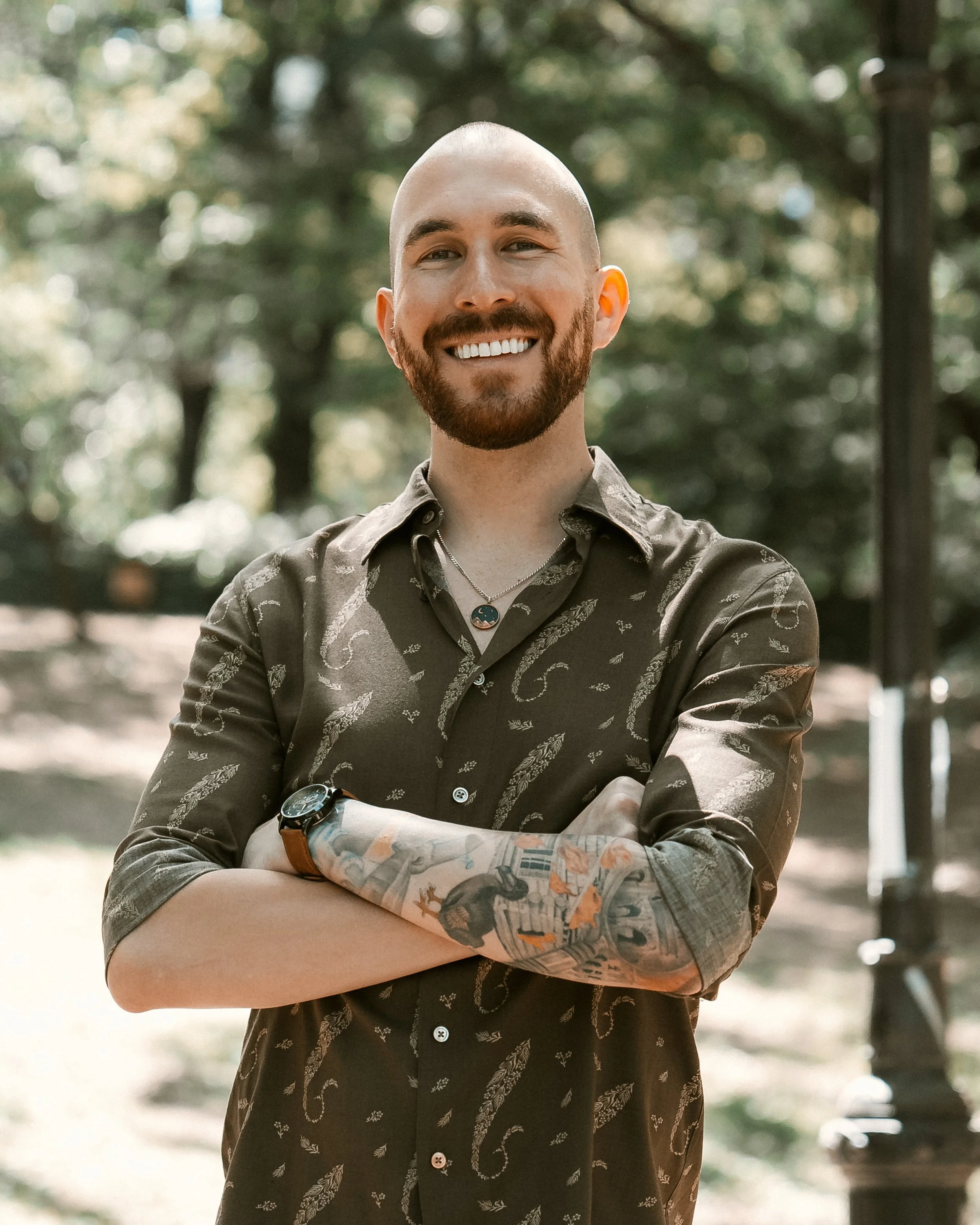 A smiling man with a beard and tattoos on his left arm, standing outdoors with trees in the background, wearing a patterned dark shirt with sleeves rolled up, a watch, and a necklace.